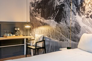 Une chambre d’hôtel comprenant un lit, un bureau avec une machine à café, une chaise et une fresque murale représentant un paysage de montagne.