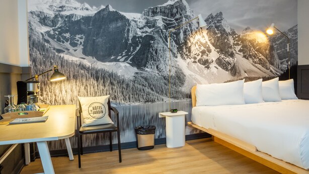 A bedroom with a large mountain mural, a desk with a lamp, a chair, and a bed with white bedding. - Hôtel du Jardin by Les Lofts Vieux-Quebec (Québec City)