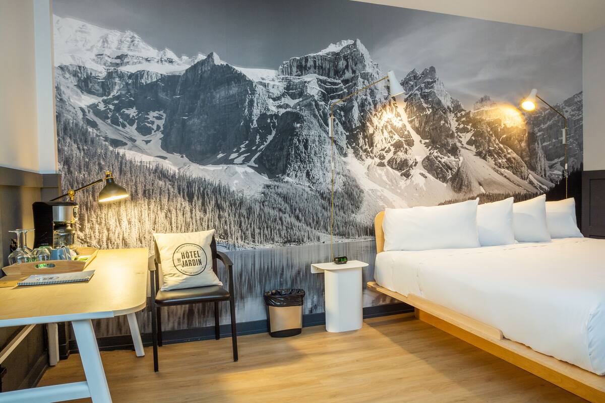 A bedroom with a large mountain mural, a desk with a lamp, a chair, and a bed with white bedding.