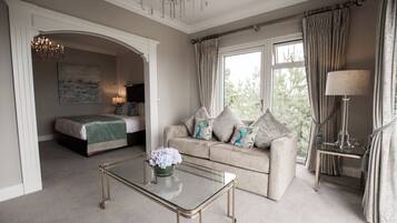 A room with a bed, a sofa, a coffee table with flowers, a lamp, and a view of greenery outside.