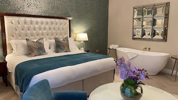 Un dormitorio con cabecera capitonada, una cama grande con cubrecama azul, una mesa redonda con un florero, una bañera y un espejo con marcos geométricos.