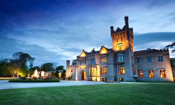 A large, historic building with a prominent tower and arched windows, surrounded by a well-maintained lawn and a body of water.