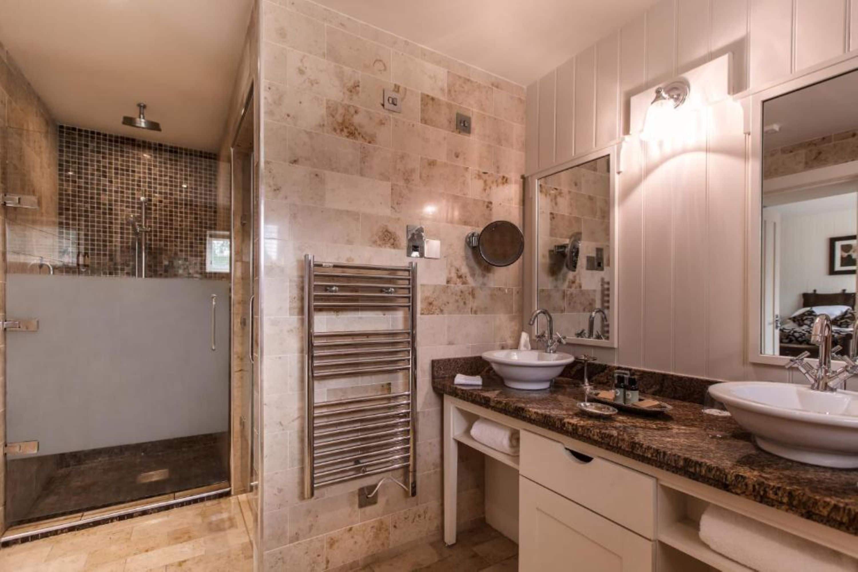 Un baño moderno con cabina de ducha de vidrio, toallero, dos lavamanos y un espejo grande.