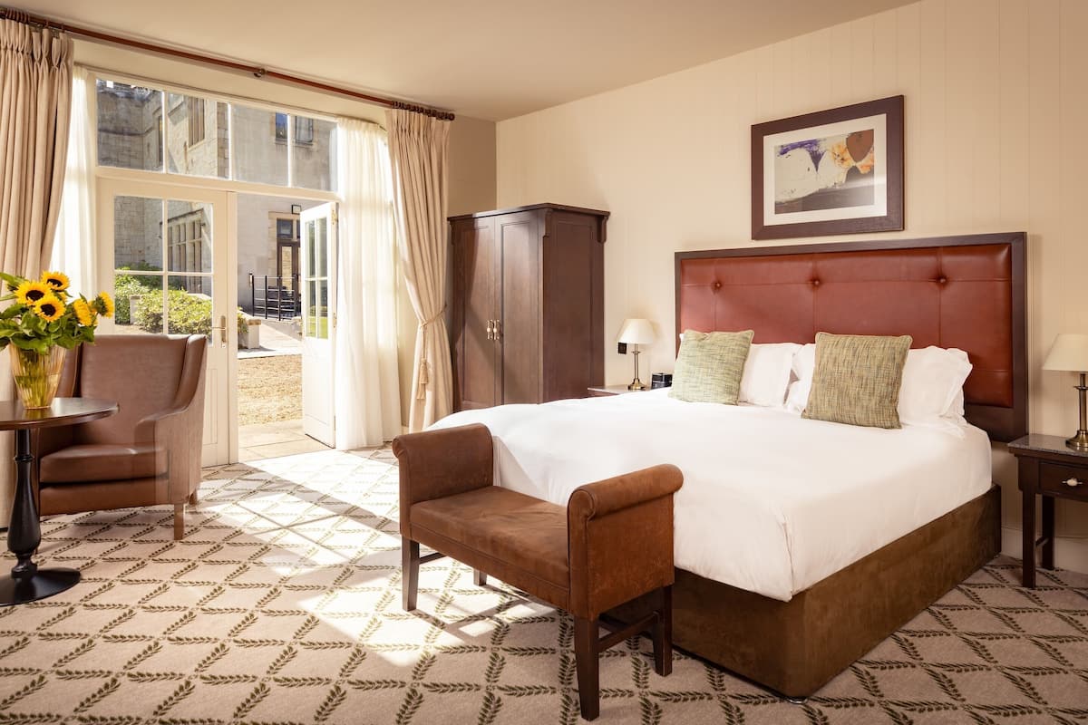 A hotel room with a large bed, a wooden wardrobe, a chair, a small table with a vase of sunflowers, and a view through a large window.