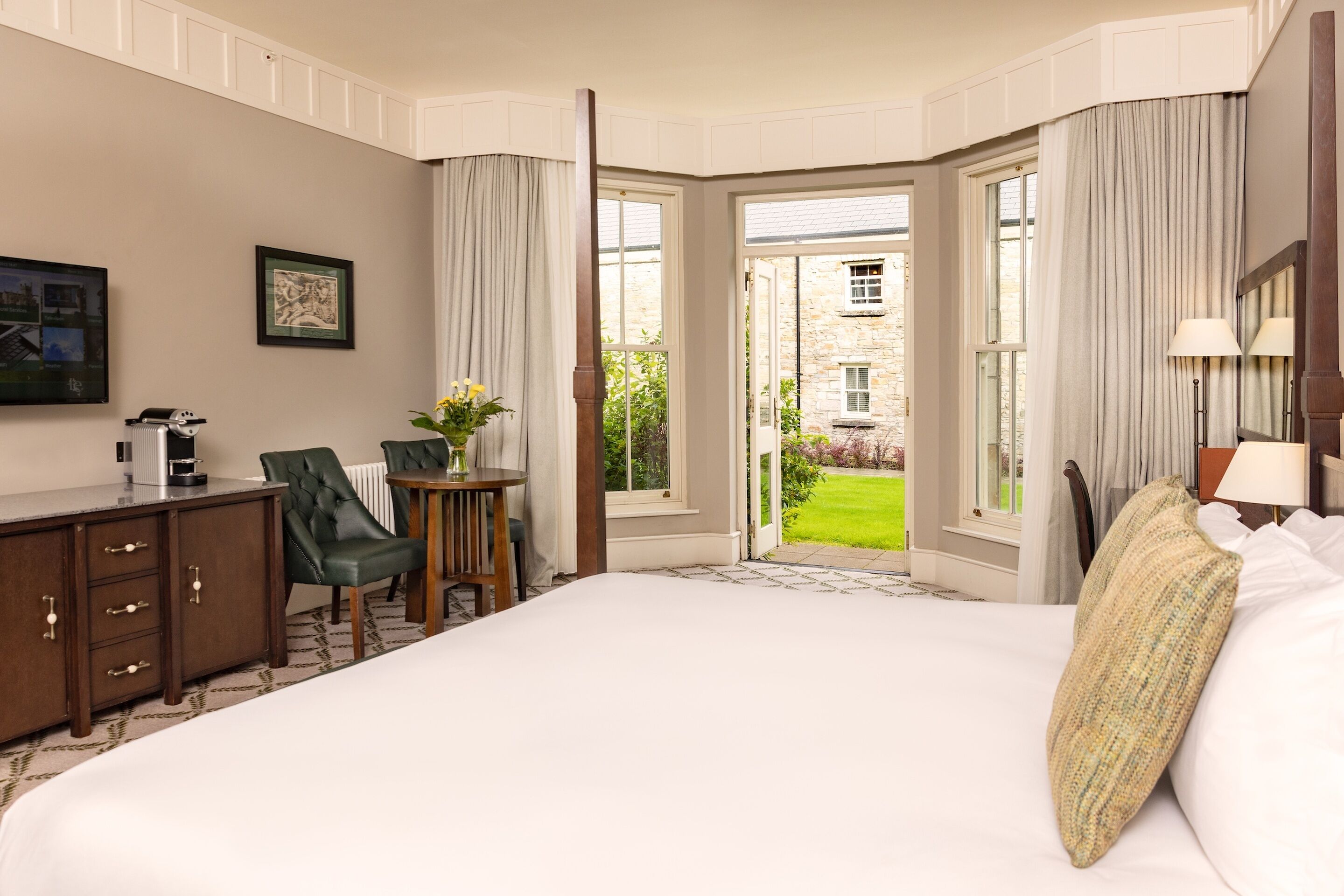 Hotelkamer met een groot bed, een bureau met stoel, een koffiezetapparaat en uitzicht op de tuin door de geopende deur.