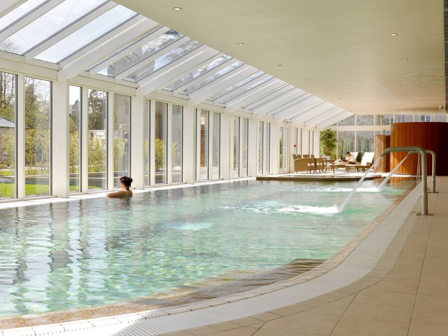 An indoor swimming pool with clear water, large windows, and a sunlit ceiling.
