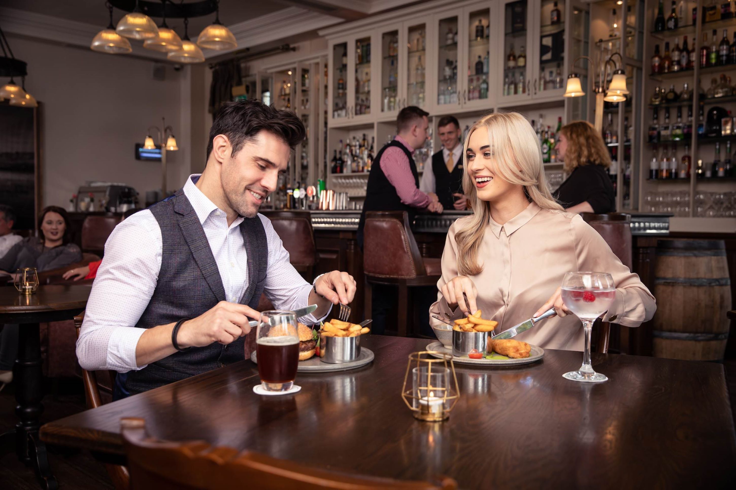 A couple dining at a bar with a variety of drinks and food items on the table.