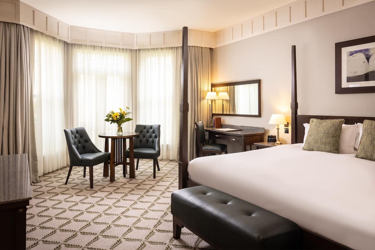 A hotel room with a large bed, a desk with a chair, a small table with flowers, and a lamp.