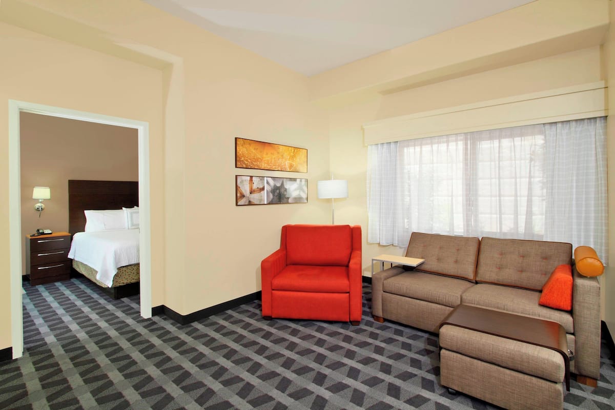 A hotel room with a bed, a red armchair, a sofa, a small table, and a window with curtains.