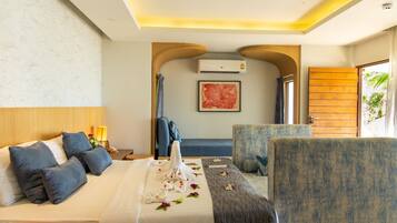 A hotel room with a bed, a sofa, two armchairs, a wooden door, an air conditioning unit, and a painting on the wall.