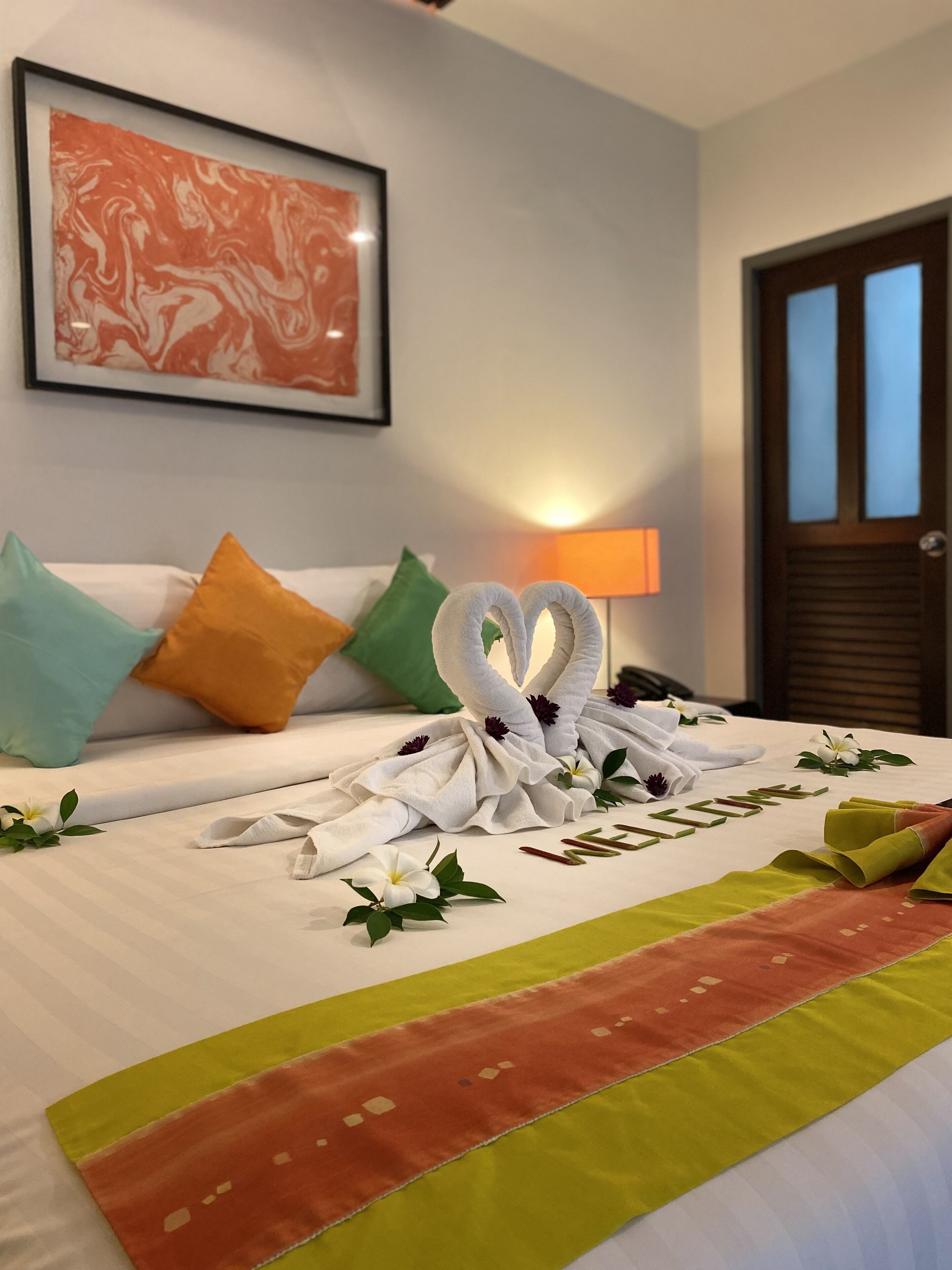 A neatly made bed with towels arranged in a swan shape, a welcome mat, and a framed artwork on the wall.
