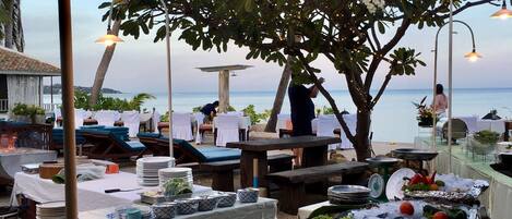An outdoor dining area with tables set for a meal, a buffet with seafood and vegetables, and a view of the ocean.