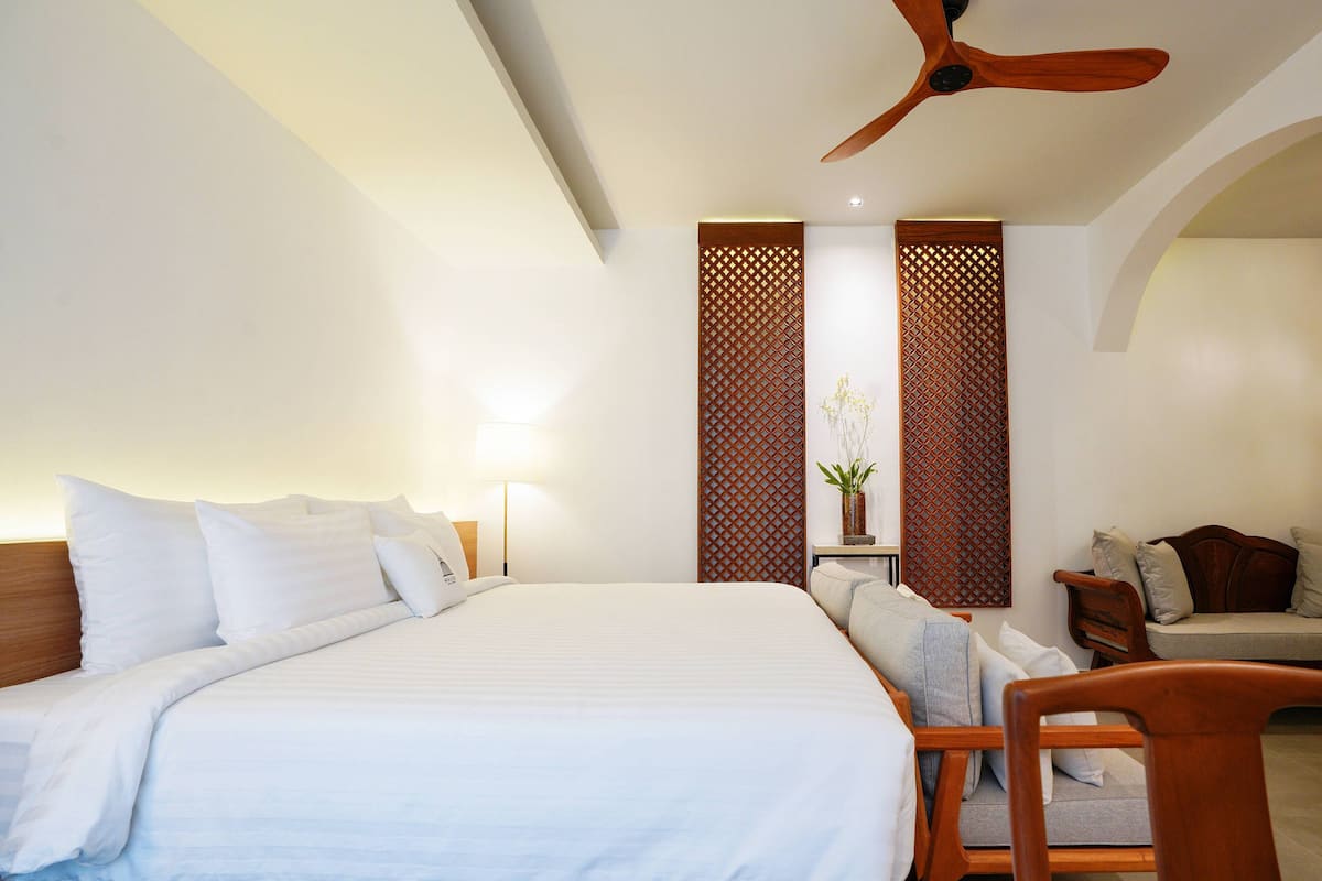 A hotel room with a large bed, a wooden ceiling fan, a small table with a vase, and a sofa.