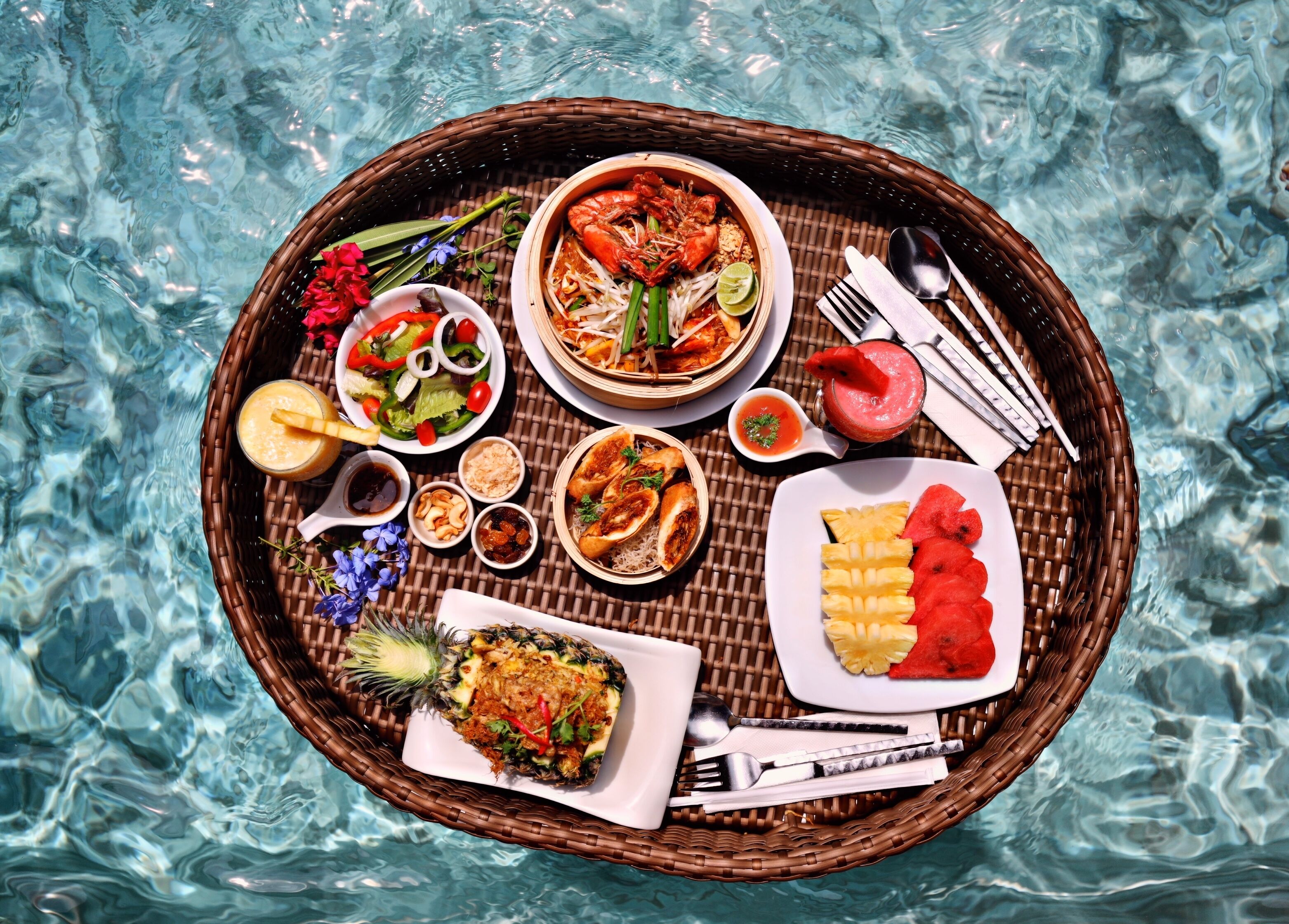 Una mesa de comedor con diversos platos, incluyendo mariscos, verduras y frutas tropicales, sobre una bandeja tejida, ubicada sobre una piscina.