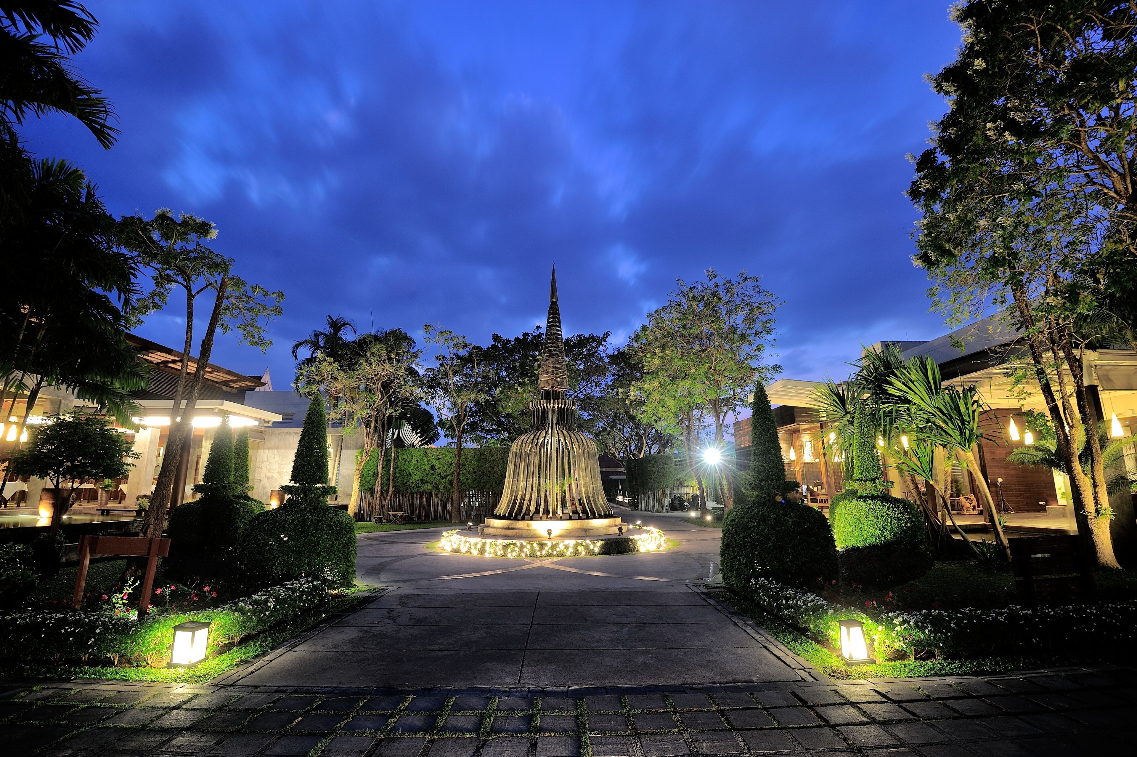 A well-lit pathway leading to a central fountain surrounded by neatly trimmed hedges and trees, with buildings on either side.