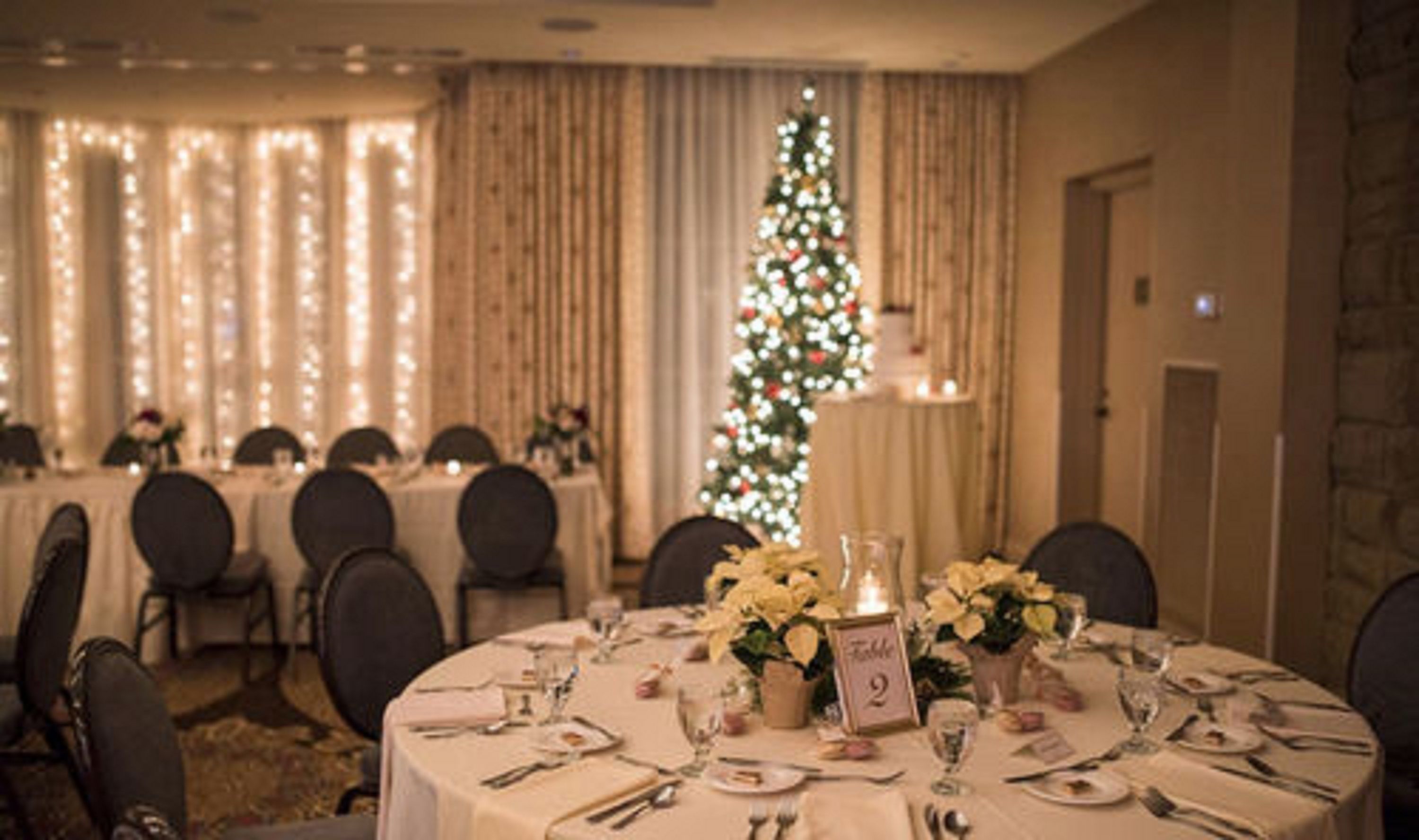 A dining area with round tables set for a meal, a Christmas tree with lights, and a warm, softly lit ambiance.