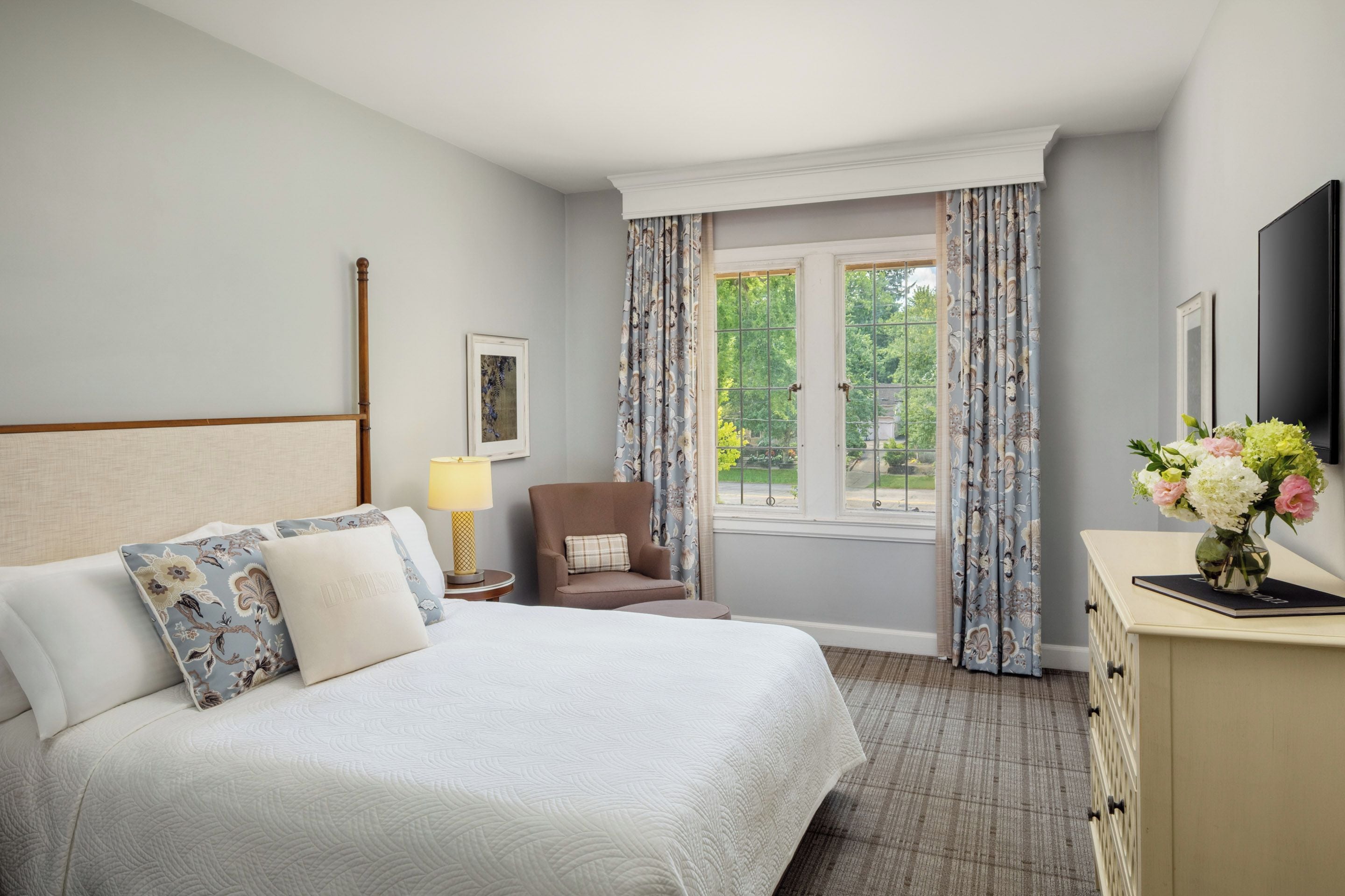A hotel room with a large bed, a chair, a desk with a vase of flowers, and a window with curtains.