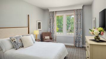 A hotel room with a large bed, a chair, a desk with a vase of flowers, and a window with curtains.