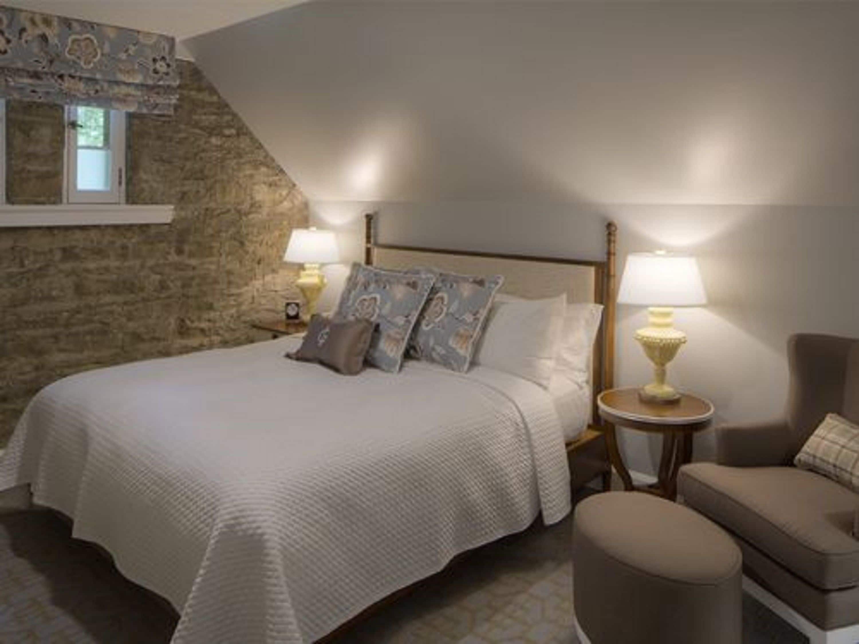 Un dormitorio con pared de piedra, una cama con ropa de cama blanca, dos lámparas de noche y una pequeña mesa redonda con una lámpara.