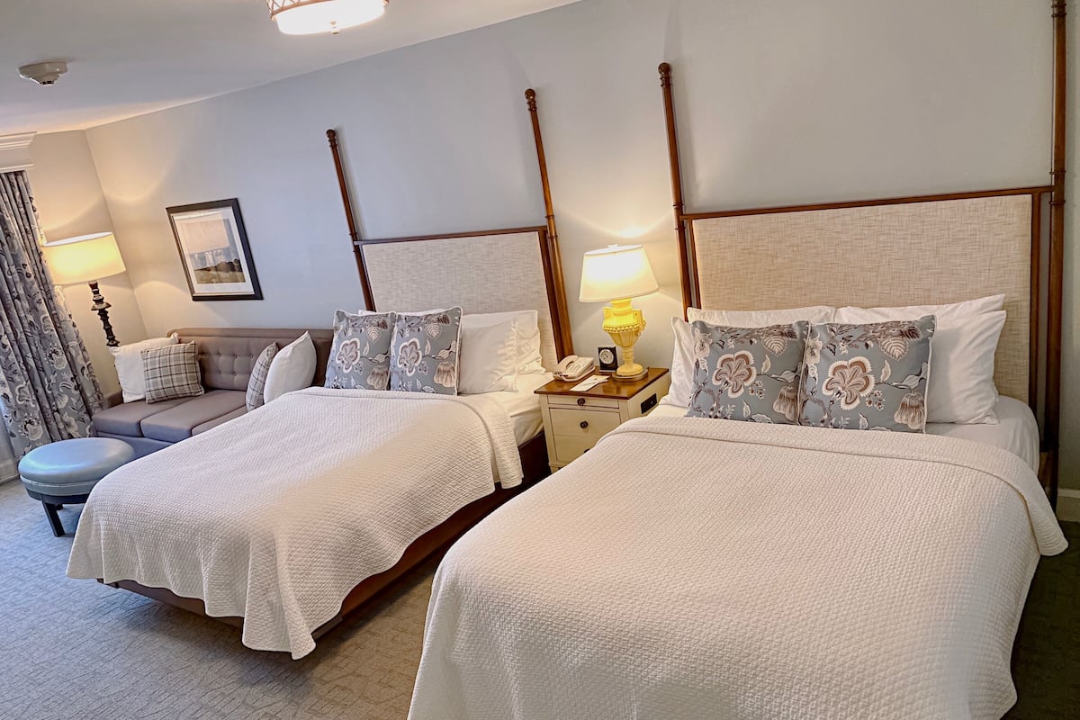 A hotel room with two beds, a sofa, a bedside table with a lamp, a telephone, and a framed picture on the wall.