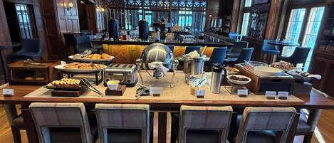 A breakfast buffet setup with various food items, a coffee machine, and seating arrangements.
