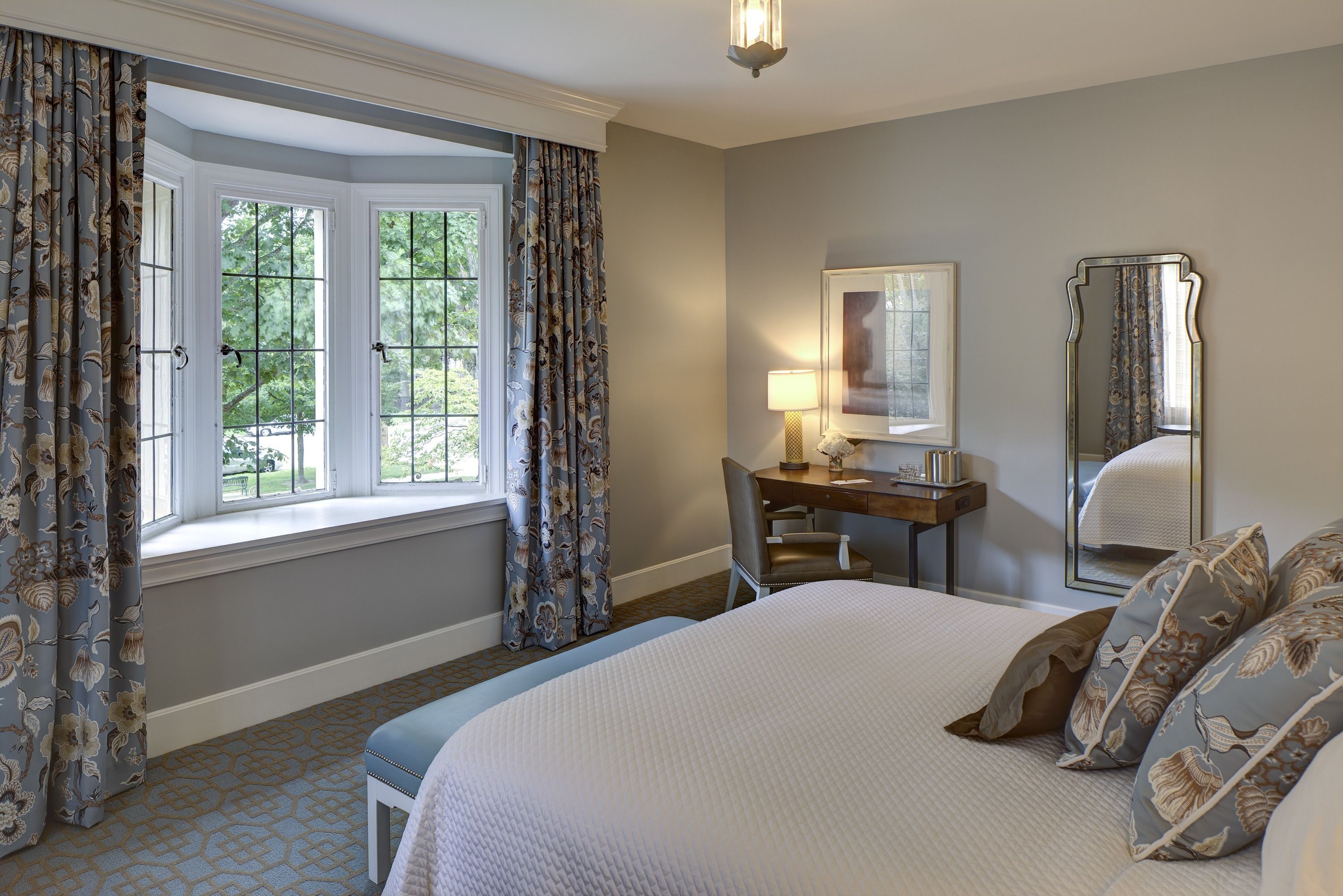 A bedroom with a large bed, a desk with a chair, a window with curtains, and a mirror.