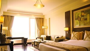 A hotel room with a large bed, a sofa, a desk, and a chair. There is a window with curtains, a painting on the wall, and a lamp on the desk. - Dia Park Premier (Gurugram)