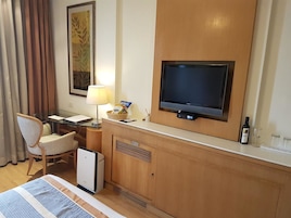 A hotel room with a bed, a desk, a television, a lamp, a bottle of wine, and a small refrigerator.
