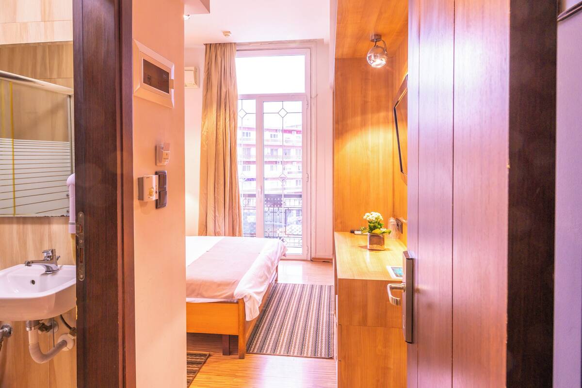 Una habitación de hotel con suelo de madera, una cama, baño con lavabo y una ventana con cortinas.