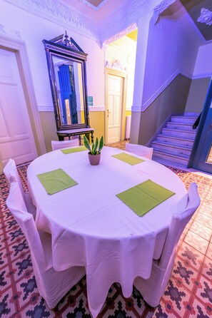 A round dining table with white tablecloth and green placemats, surrounded by white chairs, in a room with patterned floor tiles and a staircase. - Metropolis (Serres)