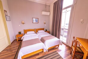 Una habitación de hotel con dos camas, un escritorio de madera, una mesita pequeña y un ventanal con cortinas.