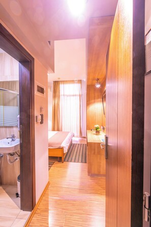 A hotel room with a wooden floor, a bed, a bedside table with a lamp, a small window with curtains, and a bathroom with a sink and mirror. - Metropolis (Serres)