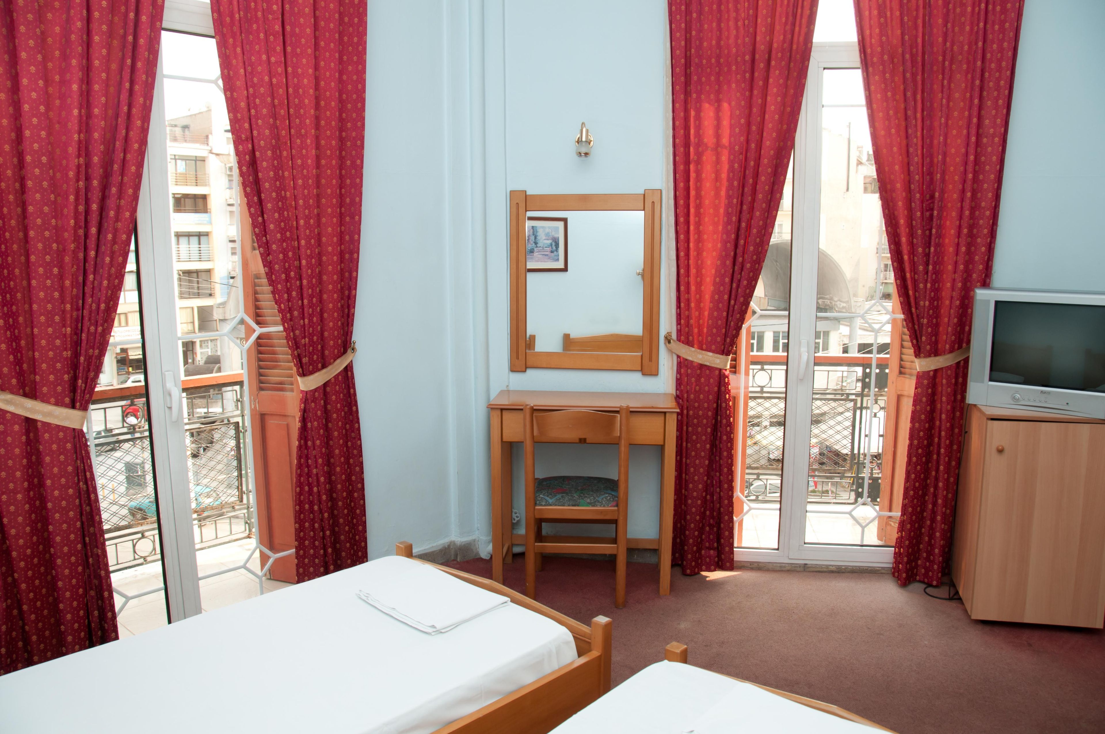 A hotel room with a bed, a desk, a chair, a television, and a window with red curtains.