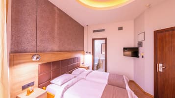 A hotel room with two beds, a wooden headboard, a bedside table with a lamp, a TV mounted on the wall, and a wooden wardrobe.