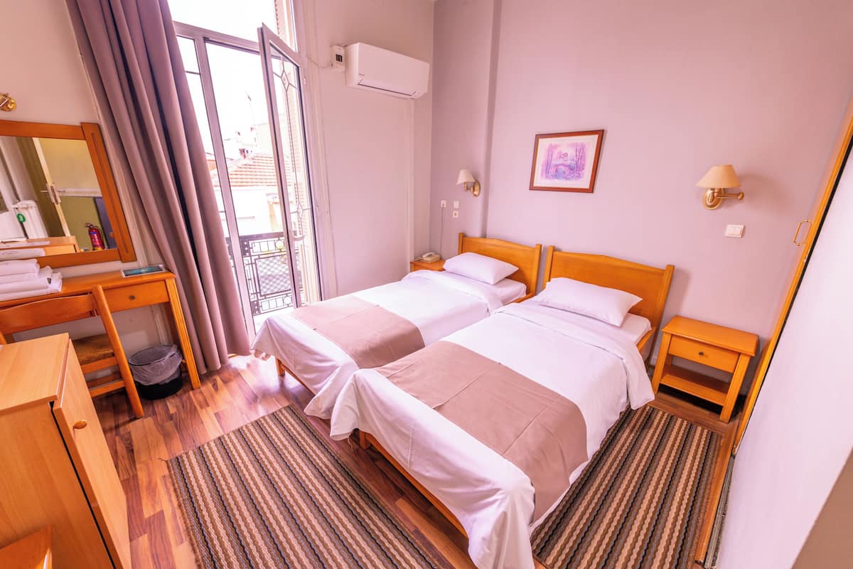 Una habitación de hotel con dos camas, un escritorio de madera, una silla, un cubo de basura pequeño, una mesita de noche con lámpara y una ventana con cortinas.