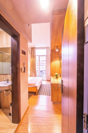 A hotel room with a wooden floor, a bed, a bathroom with a sink, and a window with curtains.