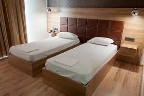 Habitación de hotel con dos camas individuales, cabeceros de madera, una mesita de noche con lámpara y una pequeña cómoda.