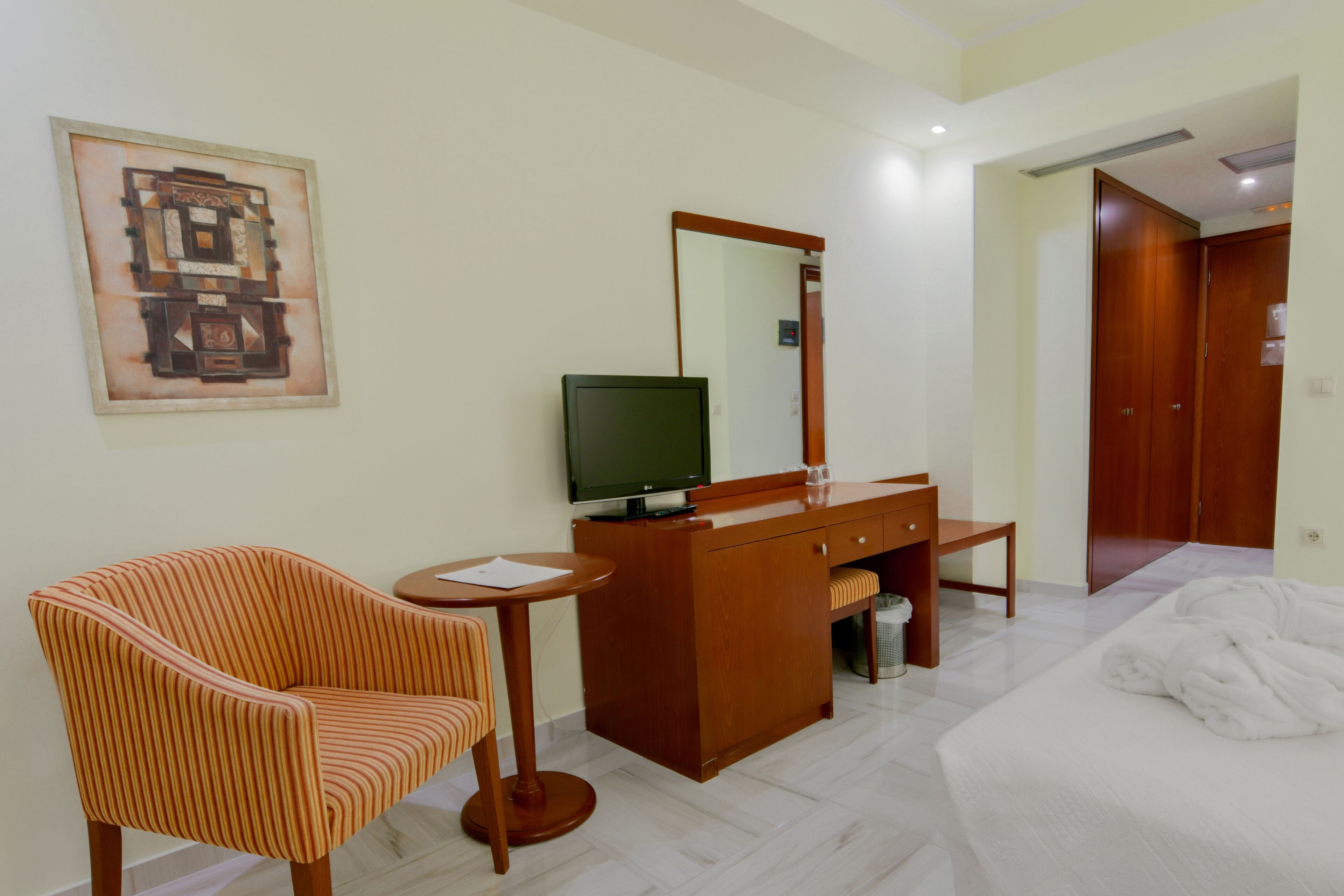 A hotel room with a bed, a wooden dresser, a television, a striped armchair, and a painting on the wall.