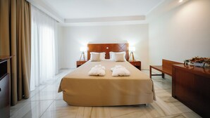 Une chambre d’hôtel avec un grand lit, une tête de lit en bois, deux tables de chevet avec des lampes, un bureau agrémenté d’un vase de fleurs et un banc.