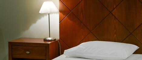 A neatly made bed with a white pillow and a white blanket, a wooden nightstand with a lamp, and a wooden headboard with a diamond pattern.