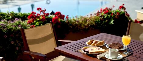 Una mesa de madera con alimentos para el desayuno, un vaso de zumo de naranja y vistas a una piscina.