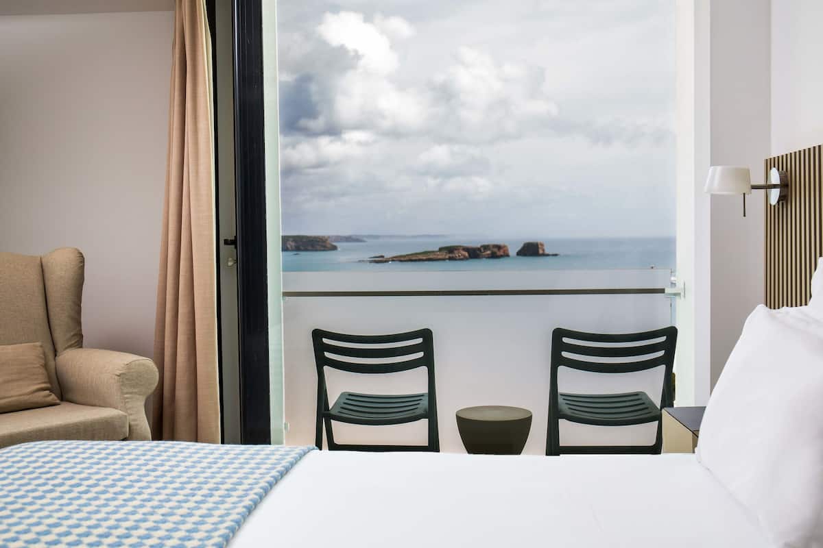 A hotel room with a bed, two chairs, a small table, and a view of the sea through a large window.