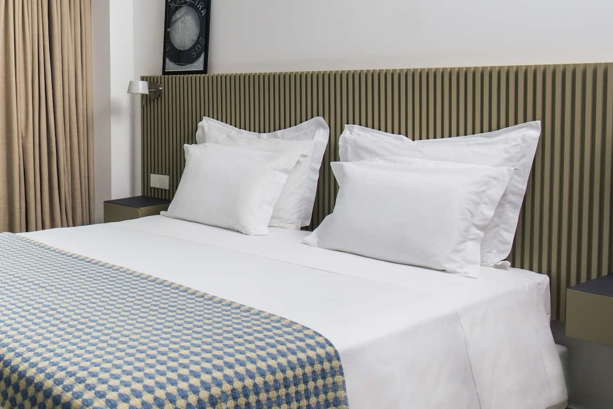 A hotel room with a bed, a checkered blanket, a headboard, a wall-mounted picture, and a window with curtains.