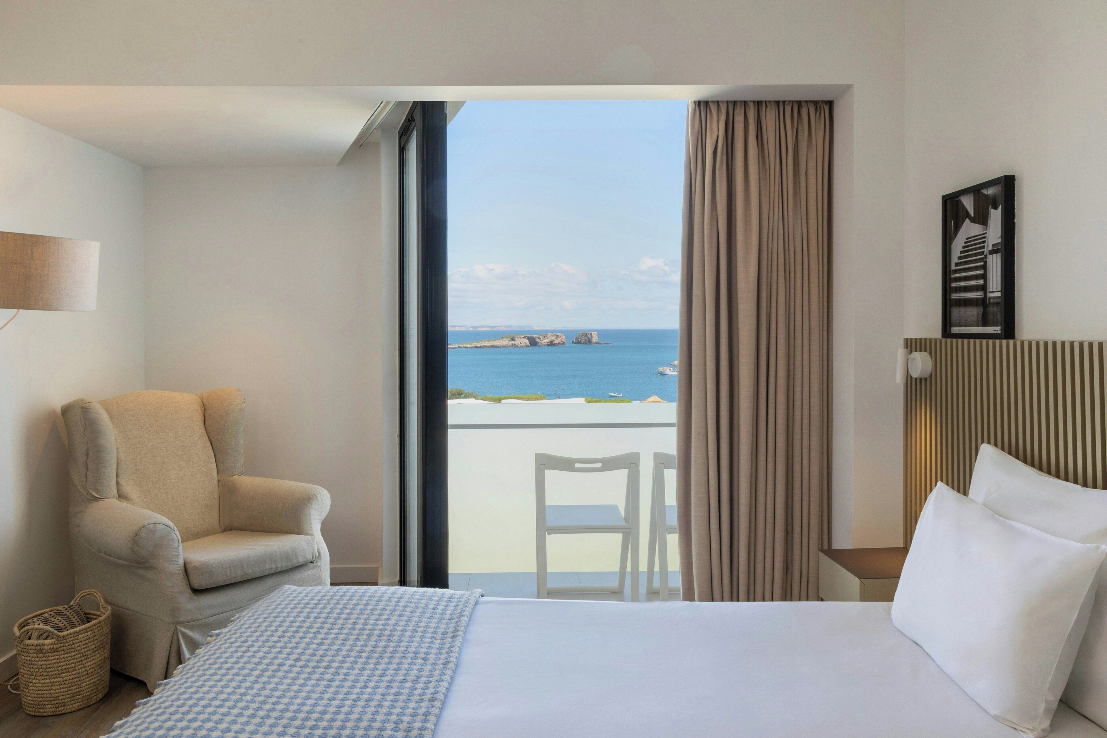 Habitación de hotel con cama, una silla, una mesita, una lámpara y vistas al mar.