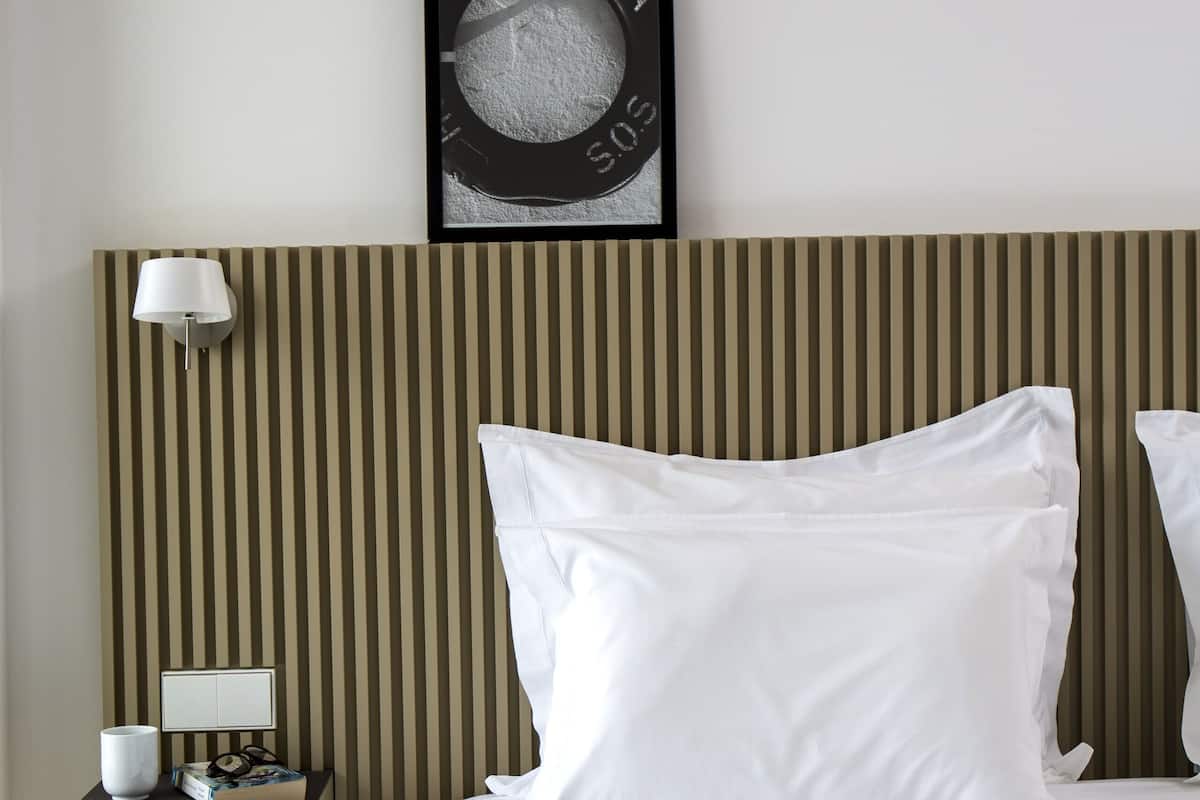 A hotel room with a striped headboard, a framed picture on the wall, a bedside lamp, a nightstand with a cup and a book, and a checkered bedspread.