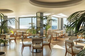 A modern indoor space with round tables and chairs, large windows, and potted plants. - Memmo Baleeira - Design Hotels (Vila do Bispo)