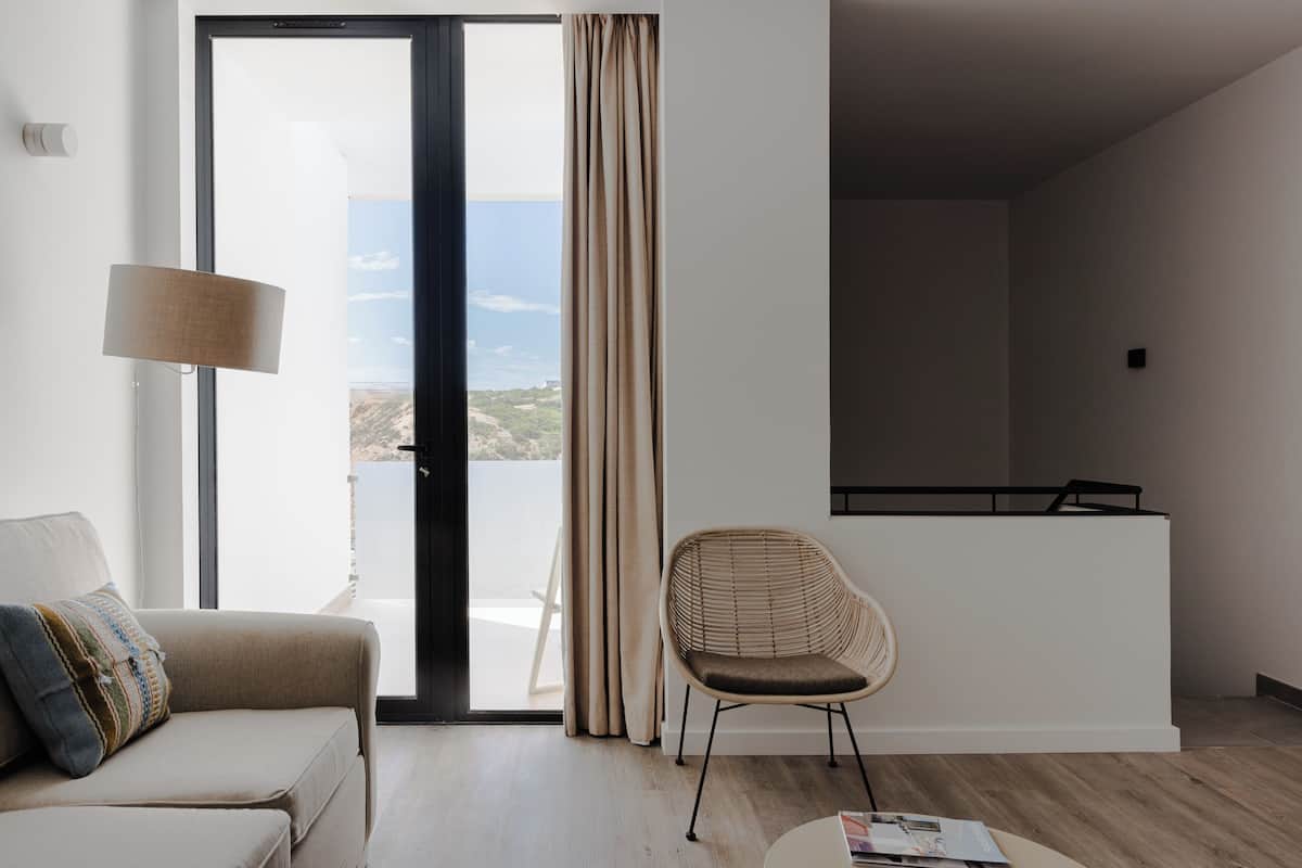 A modern hotel room with a sofa, a wicker chair, a small table, and a view of the sea through a large window.