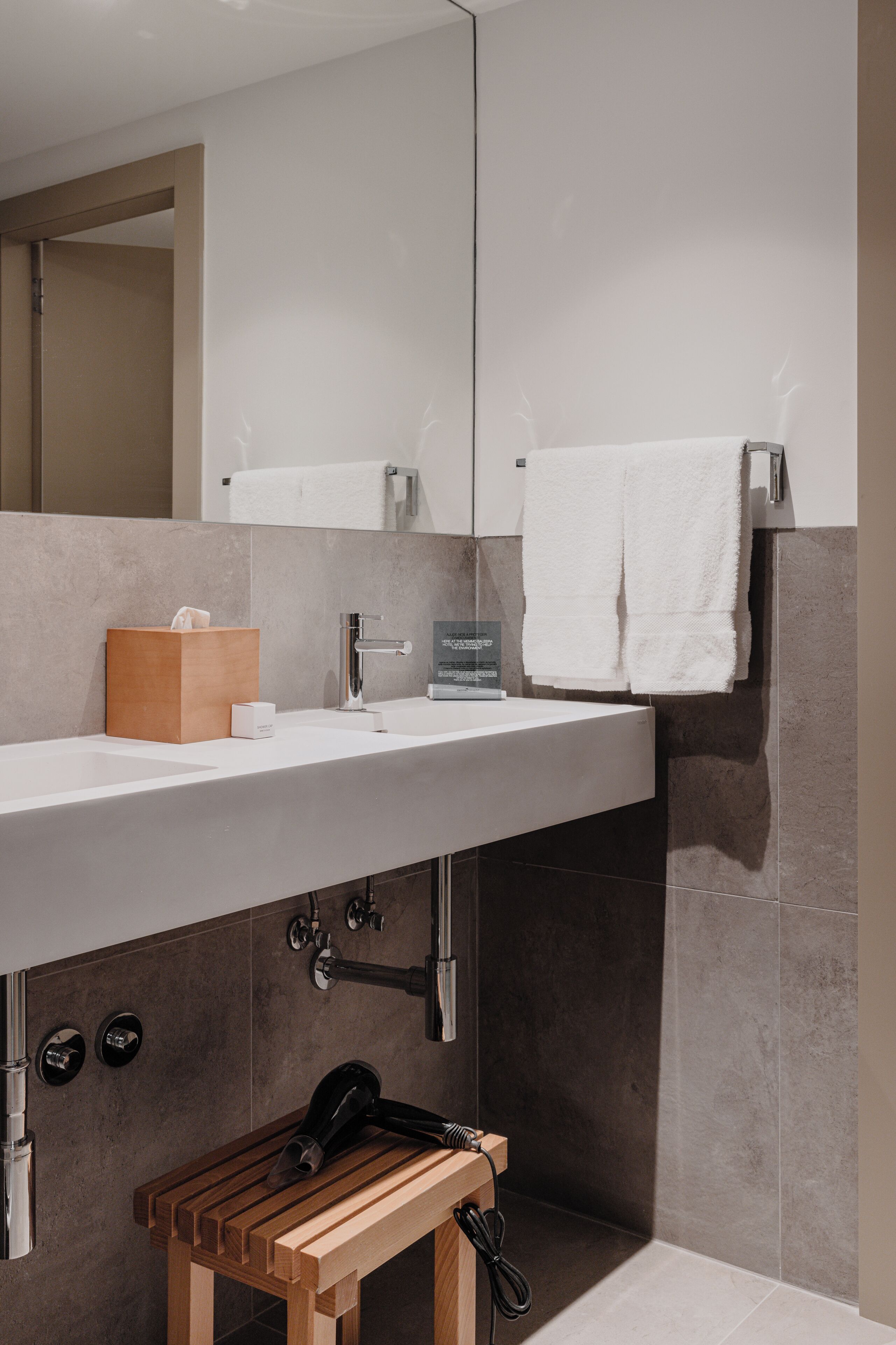 A modern bathroom with a large mirror, a white sink, a wooden bench, and a towel rack.
