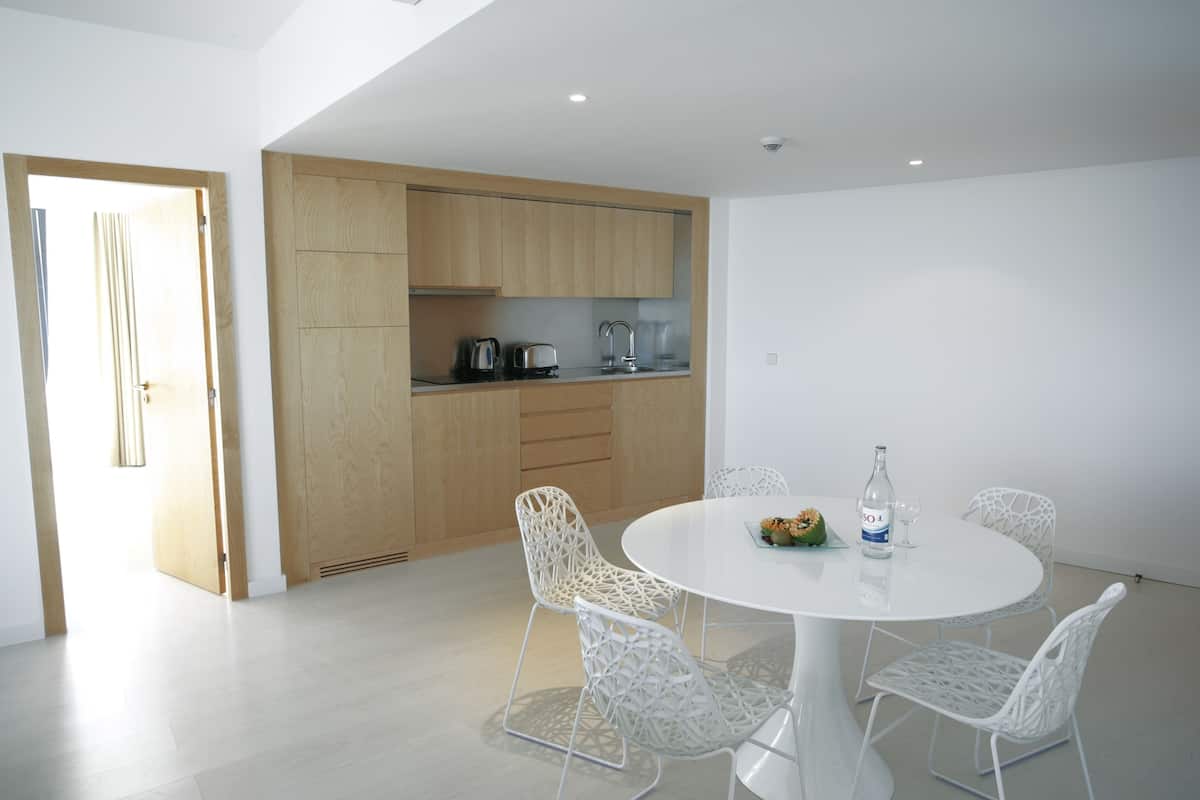 A modern kitchen with wooden cabinets and a white countertop. There is a round dining table with white chairs and a bottle of water.