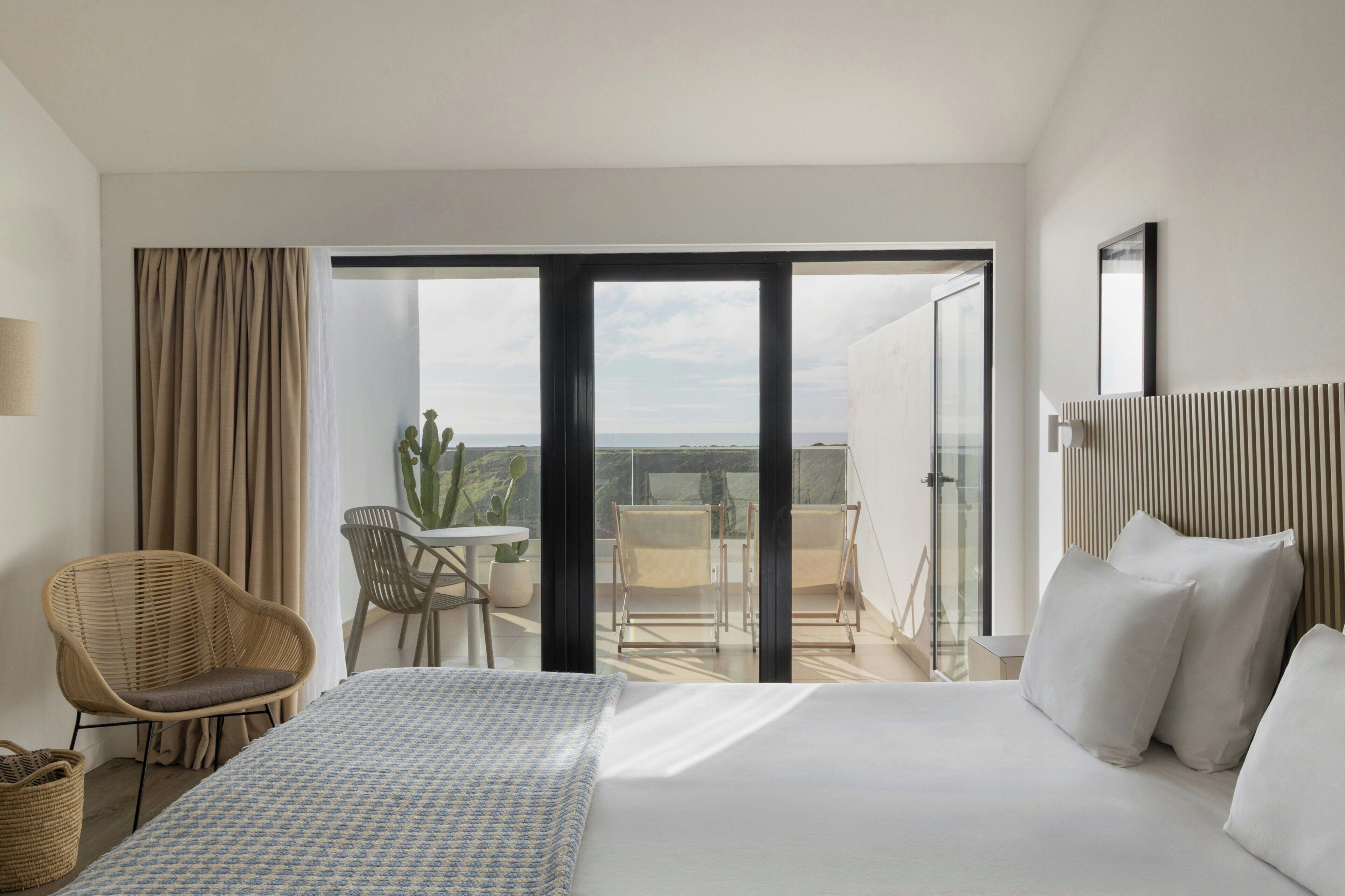 A hotel room with a bed, a chair, a table, and a view of the outdoors.